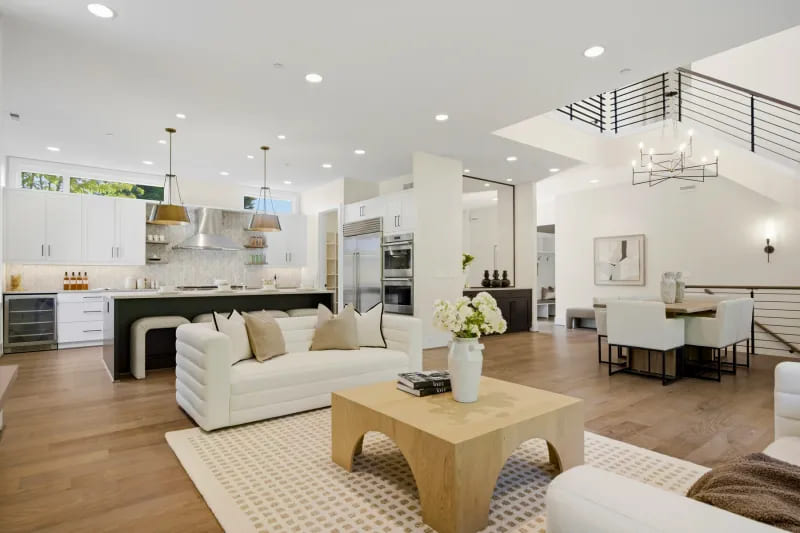 Open living, dining, and kitchen area with pale oak floors, a white sofa, an island with pendant lights, and an open staircase.