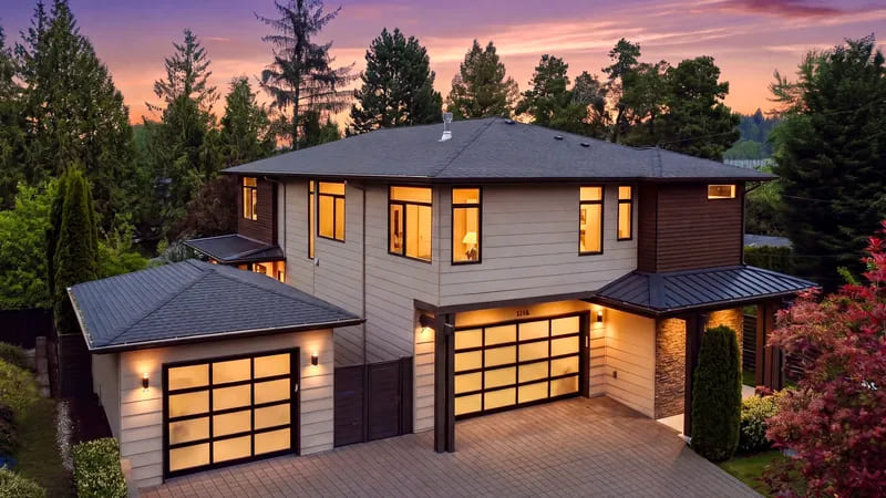 Contemporary two-storey home at twilight with lit windows, glazed garage doors, and a brick driveway beneath a violet sky.