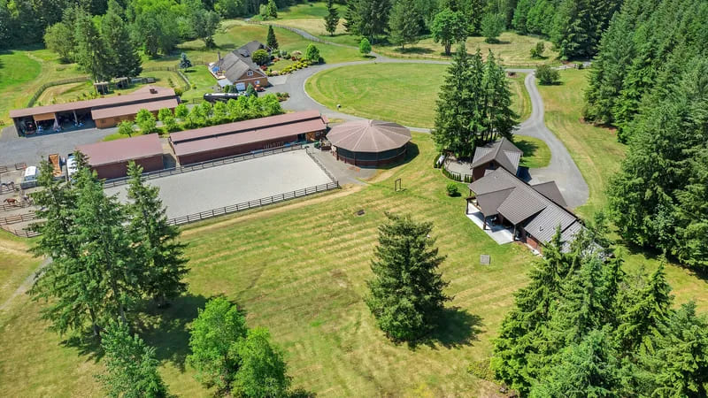 Aerial view of a long barn, a covered round pen, and a sand riding arena beside pasture and evergreen forest, with the home on the far side of the property.