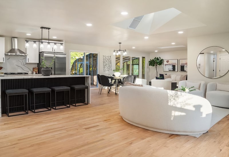 Open kitchen with a dark island and bar stools, looking through to a curved sofa in the living area and a dining table beyond.