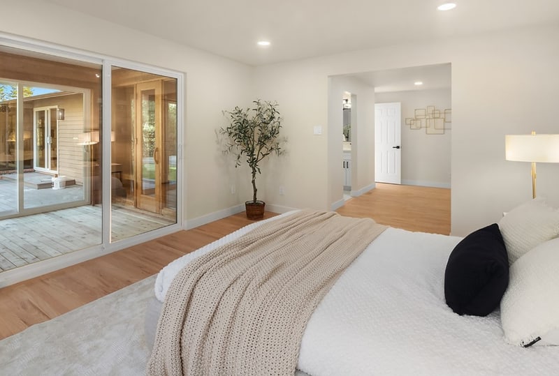 Bedroom with a low bed and a floor lamp, opening through sliding glass doors to an adjoining room with a potted olive tree.