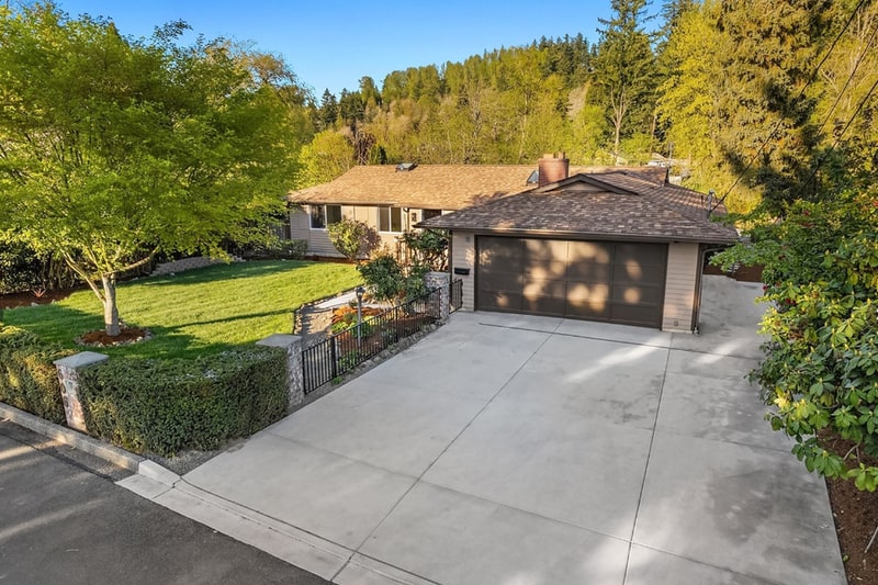 Single-storey home with a brick chimney and a wide concrete drive, set behind a lawn, a clipped hedge, and mature trees on a hillside lot.