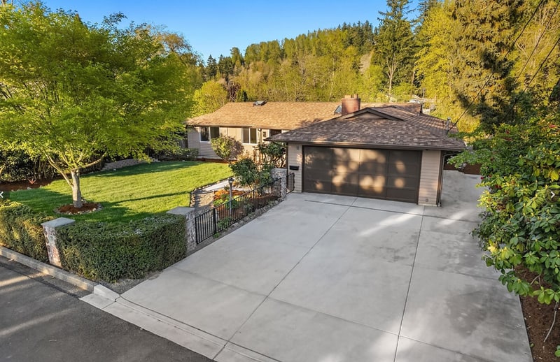 Single-storey home with a brick chimney and a wide concrete drive, set behind a lawn, a clipped hedge, and mature trees on a hillside lot.