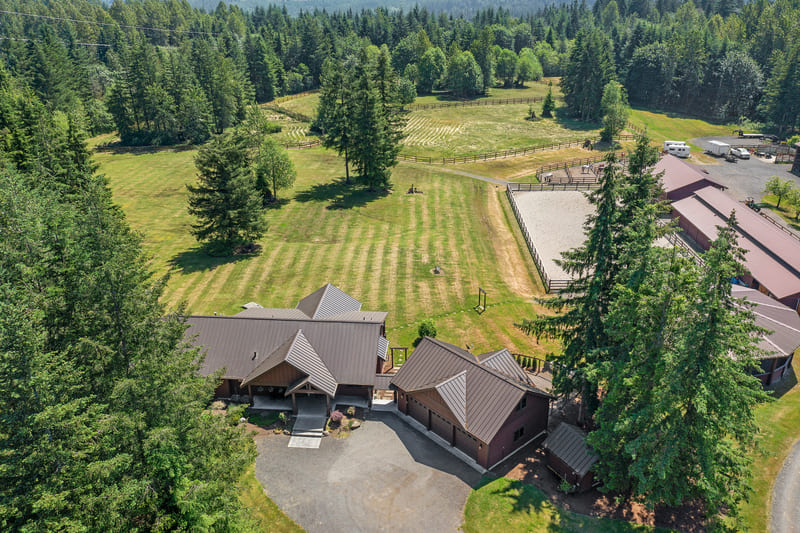 Aerial view of a timber and metal-roofed home with a circular gravel drive, mown pasture, a sand riding arena, and a long barn set among evergreen trees.