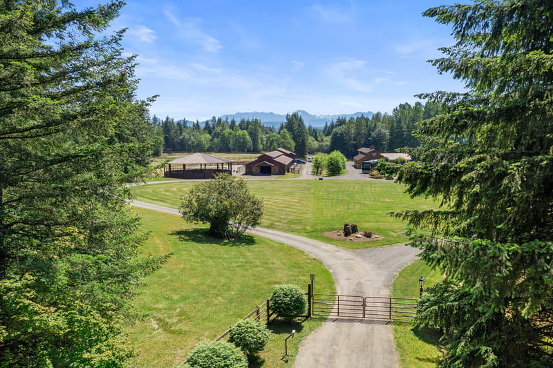 Aerial view from a gated gravel entrance looking across lawn and pasture toward the barn, riding arena, and distant foothills.