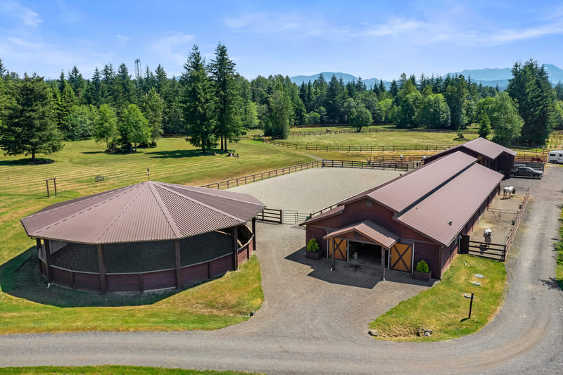 Aerial view of a covered round pen beside a long barn with a centre breezeway and an adjoining sand riding arena.