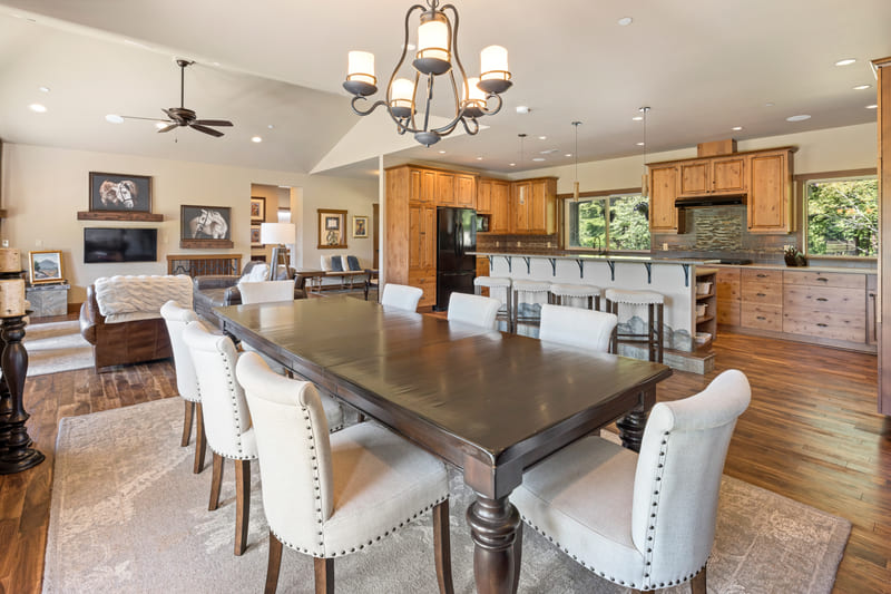 Great room with a long dining table beneath a wrought-iron chandelier, open to the kitchen and a living area with a stone fireplace.