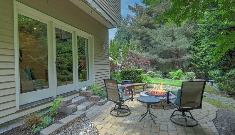 Paved back patio with a table, chairs, and a fire bowl beside tall windows, looking out to a lawn edged with evergreens.