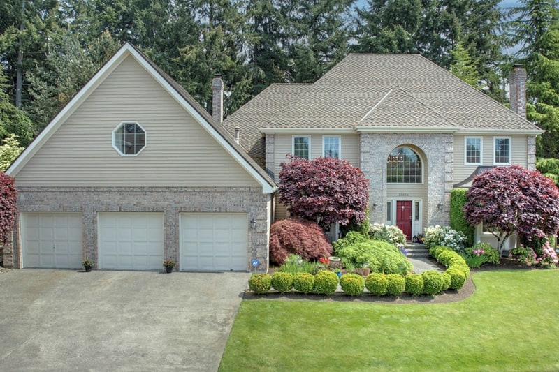 Two-storey home with pale siding and a stone-faced arched entry, a three-car garage, and red maples and clipped shrubs along the front lawn.