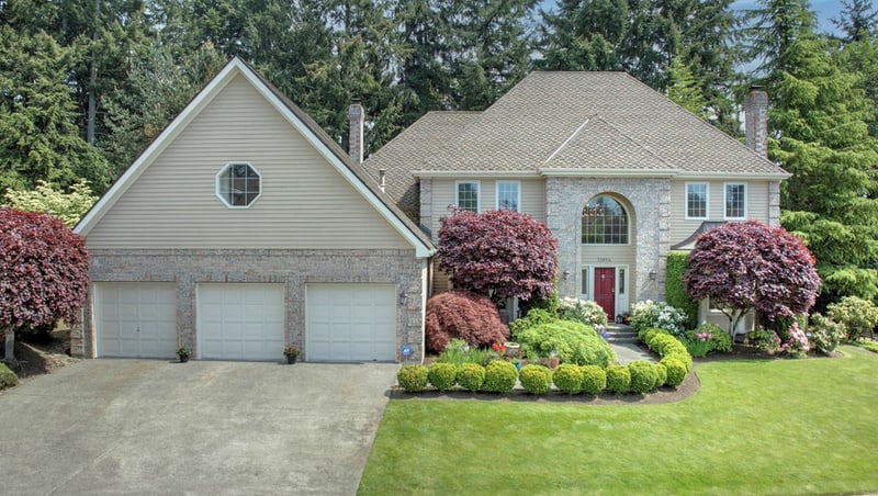 Two-storey home with pale siding and a stone-faced arched entry, a three-car garage, and red maples and clipped shrubs along the front lawn.
