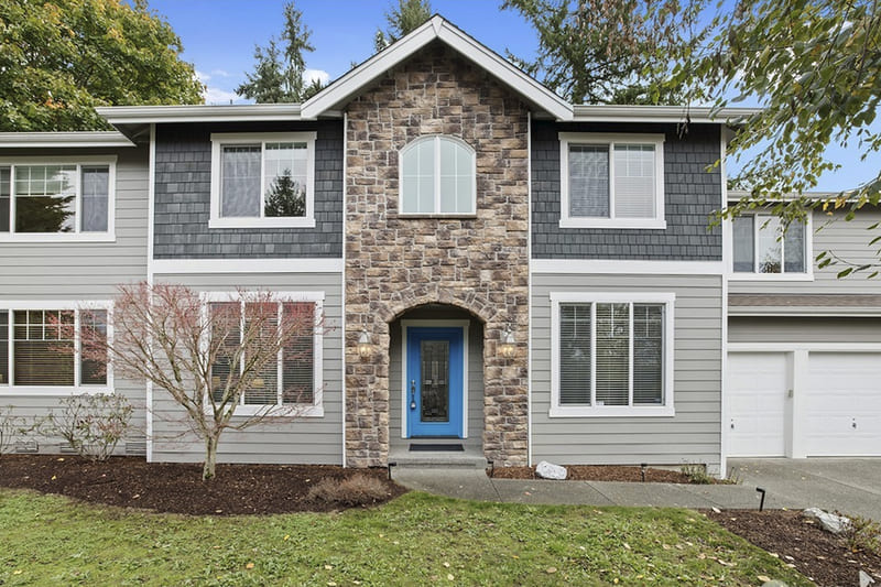 Two-storey home with grey siding, shingle accents, a stone-faced entry arch, a blue front door, and a lawn bordered by planting beds.