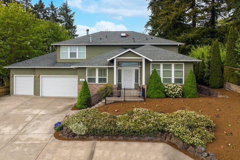 Two-storey home with a gabled dormer and a covered entry, a two-car garage, and a curved planting bed beside a concrete drive, with evergreens behind.