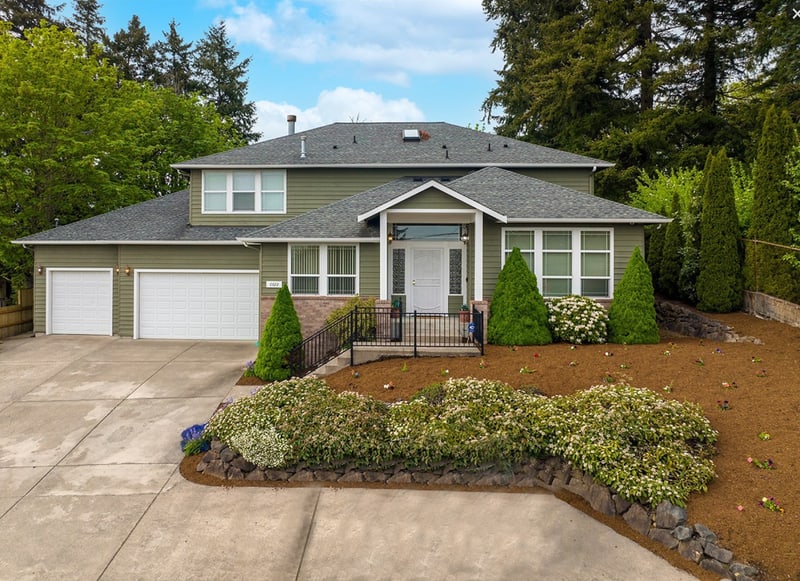 Two-storey home with a gabled dormer and a covered entry, a two-car garage, and a curved planting bed beside a concrete drive, with evergreens behind.