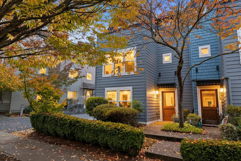 Blue-sided townhome at dusk with a covered wood front door, trimmed hedges, and stone steps, framed by a maple in autumn colour.