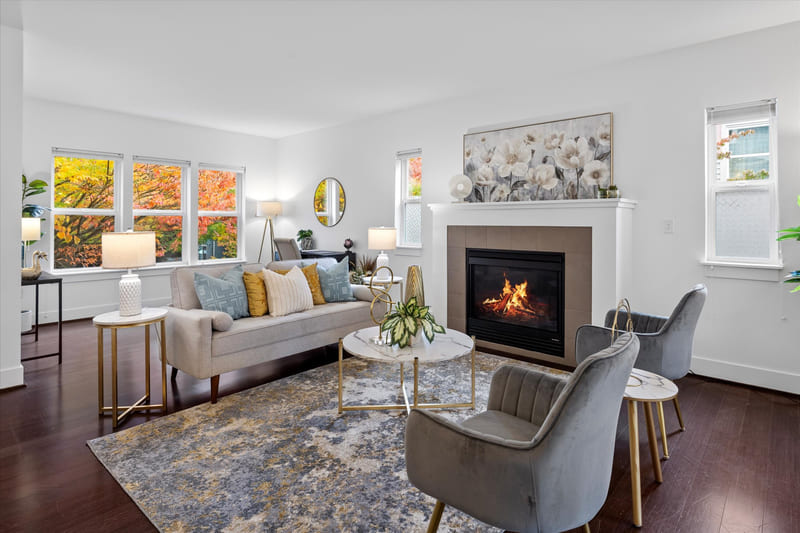 Living room with a tiled gas fireplace, a light sofa with patterned cushions, two grey armchairs, and windows showing autumn foliage.