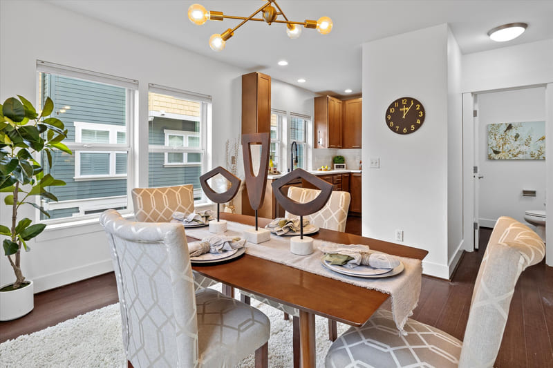 Dining area with a wood table set for four, upholstered chairs, a brass light fixture, and an open kitchen beyond.